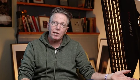 Man in gray zip-up hoodie and glasses speaking during an interview or presentation in an office setting.
