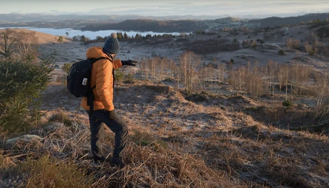 Hiker in orange jacket with backpack overlooking a valley with lakes and bare trees during golden hour.