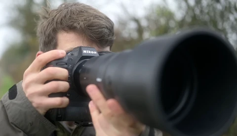 Photographer holding a Nikon DSLR with telephoto lens aimed at camera. Blurred background shows trees and foliage.