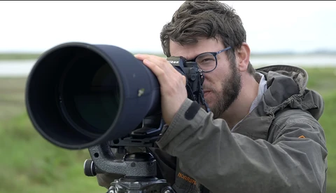 Photographer with glasses looking through a large telephoto lens mounted on a tripod in an open field.