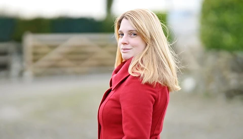 Woman with blonde hair wearing a red jacket, looking over her shoulder outdoors.