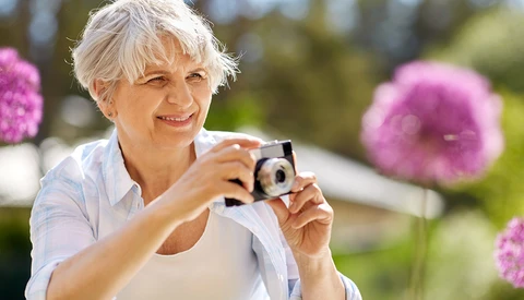 Older woman with short white hair holding a compact digital camera in a garden with pink flowers.