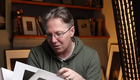 Man wearing glasses and olive hoodie reviewing contact sheets at a desk.