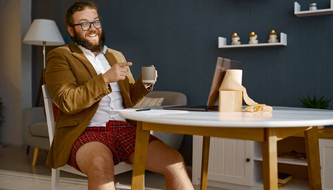 Man in mustard blazer and red plaid shorts sitting at desk with coffee cup and laptop.