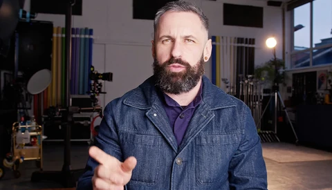 Bearded man in denim shirt gesturing while speaking in a professional studio setting.