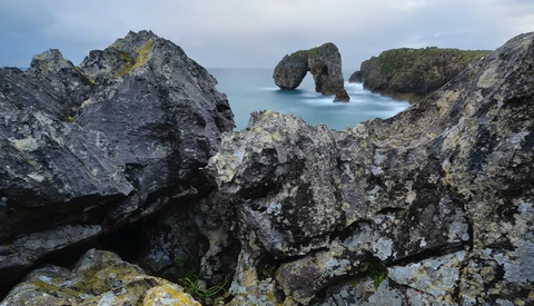 Rugged coastal rock formations with turquoise water and a natural stone arch in the distance.