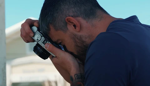 Photographer holding a mirrorless camera to his face in bright daylight conditions.