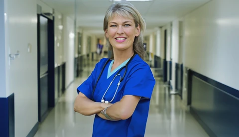 Healthcare professional in blue scrubs with stethoscope, arms crossed in hospital corridor.