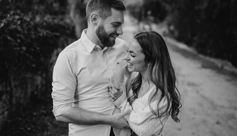 Couple embracing and smiling outdoors on a tree-lined path in black and white.