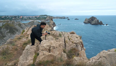 Photographer crouching on rocky coastal outcrop while framing a shot of distant sea stacks and turquoise waters.