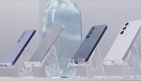 Five smartphones in different colors displayed at angles on clear acrylic stands with a translucent blue bottle in the center background.