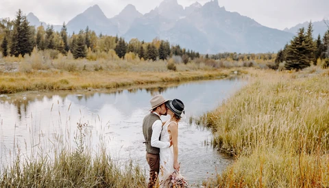Couple wading in shallow river surrounded by golden marsh grasses with forested mountains in the background.