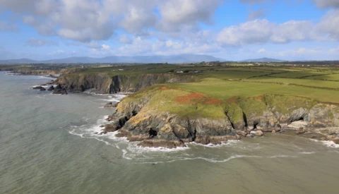 Coastal headland with steep green cliffs dropping to rocky shoreline and churning surf below.