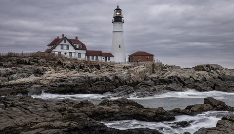 White lighthouse with red-roofed keeper's house on rocky Maine coast during overcast conditions.