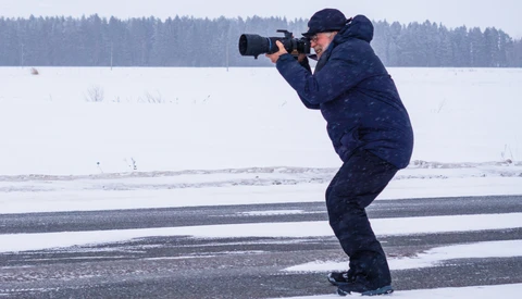 Photographer in dark winter coat crouched on frozen lake while shooting with telephoto lens, forest treeline visible across snowy landscape.