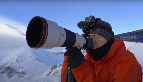 Photographer with telephoto lens shooting from aircraft window above snowy mountain landscape.