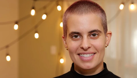Portrait of a young person with short reddish-brown hair wearing a black turtleneck, smiling at the camera against a warm-lit background with bokeh lights.