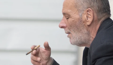 Man in profile holding a cigar, wearing dark clothing against a blurred white background.