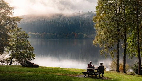 Two people sitting at a picnic table on a grassy shore, facing a calm lake surrounded by evergreen forest under misty mountains.