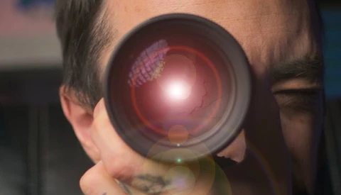 Close-up of a photographer's face looking through a camera lens with a bright red glow and internal reflections visible.
