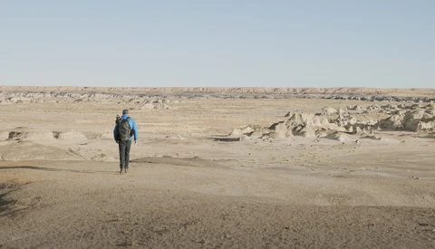 Solitary hiker with blue backpack walking across vast, barren desert terrain scattered with rocks.