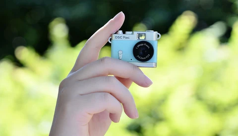Hand holding a light blue vintage compact camera against a blurred green foliage background.