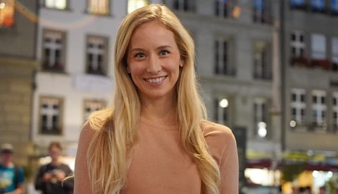Blonde woman smiling at camera in front of illuminated European buildings at dusk.