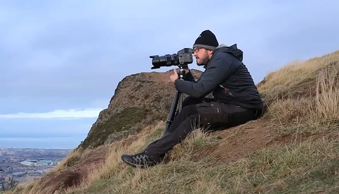 Photographer in dark jacket crouched on hillside aiming telephoto lens toward distant landscape.