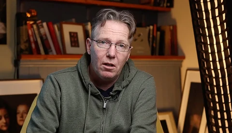 Man wearing glasses and olive hoodie speaking in front of bookshelf.