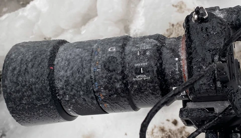 Telephoto lens covered in frost and ice mounted on camera body in snowy conditions.