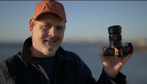 Photographer holding a mirrorless camera with telephoto lens against a blurred waterfront backdrop.