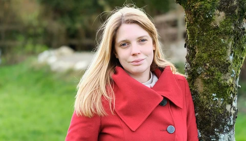 Young woman with blonde hair wearing a red coat, leaning against a moss-covered tree in an outdoor setting.