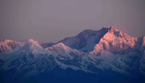 Snow-covered mountain range illuminated by alpenglow at sunrise or sunset, with pink and warm light on peaks against a cool blue sky.