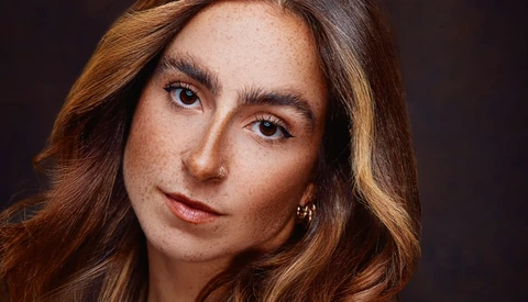 Portrait of a woman with long reddish-brown hair and freckles against a dark background.