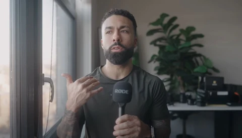 Male content creator speaking into a microphone while gesturing toward a window in a studio setting.