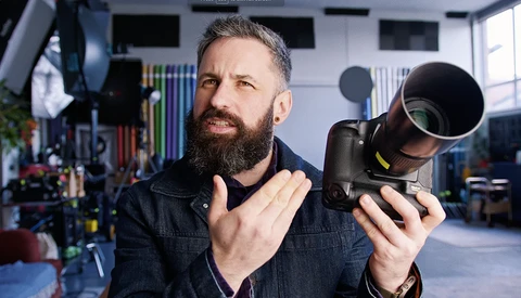 Bearded photographer holding a telephoto lens in a studio setting, demonstrating camera equipment.