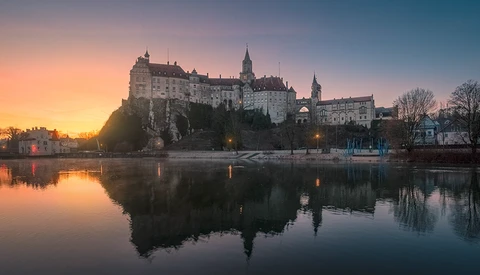 Medieval castle with multiple spires reflected in still river water at sunrise.