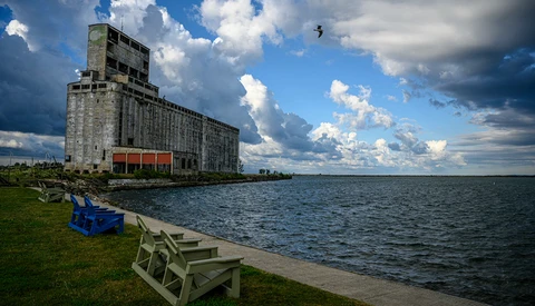Abandoned grain elevator tower beside a calm waterfront with Adirondack chairs on the grass.