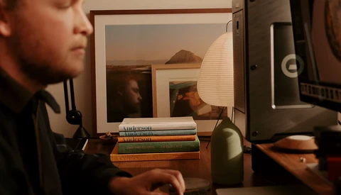 Desk setup with stacked books, desk lamp, and monitor displaying interior photographs.