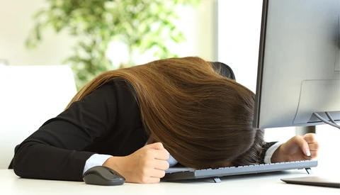 Person in black blazer slumped over desk with head resting on keyboard.