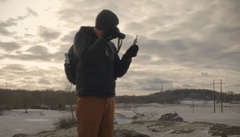 Photographer silhouetted against overcast sky while shooting by a frozen waterway with utility poles visible on distant shore.