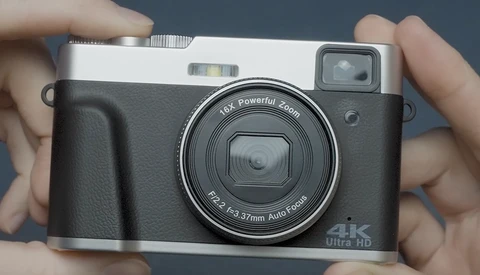 Compact digital camera with black body and silver top plate held in hands against gray background.