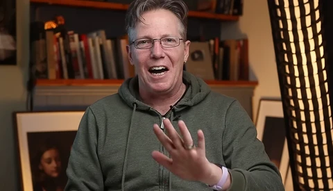 Man in glasses and olive hoodie gesturing while speaking, with bookshelves and warm lighting in background.