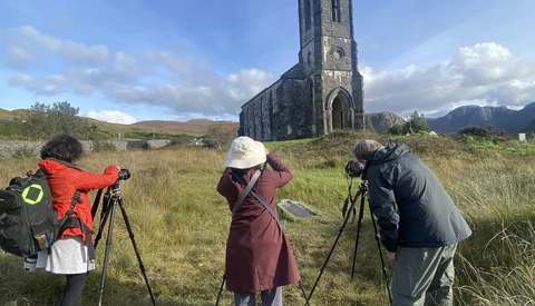 Three photographers with tripod-mounted telephoto lenses facing a stone church ruin in a mountain valley.