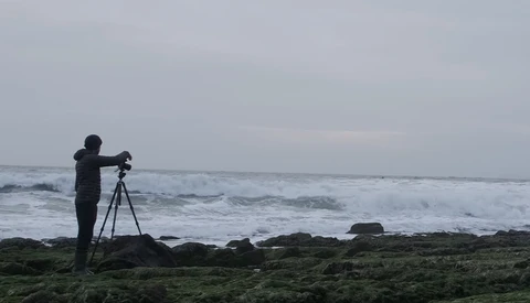 Photographer on tripod positioning equipment on rocky shore facing turbulent ocean waves.
