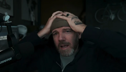 Man with beard and tattoos shading his eyes while speaking into a studio microphone.