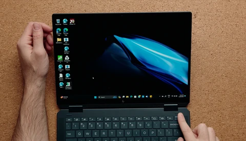 Hands holding a laptop displaying Windows desktop with blue abstract wallpaper and desktop icons visible on left sidebar.