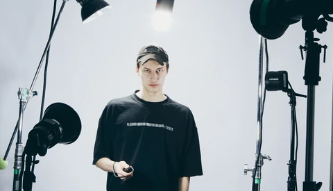 Man in black t-shirt and cap stands in professional studio surrounded by lighting equipment.