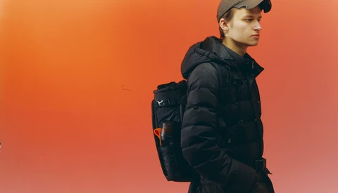 Young man in black puffer jacket and cap standing against warm orange gradient background.