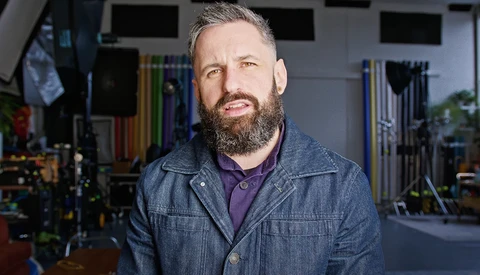Bearded man with gray hair wearing denim jacket over purple shirt in workshop studio.